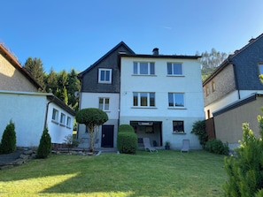 Un immeuble résidentiel à plusieurs étages, doté d’un toit plat, d’une entrée bien visible et d’un petit jardin devant, avec de la pelouse et deux chaises.