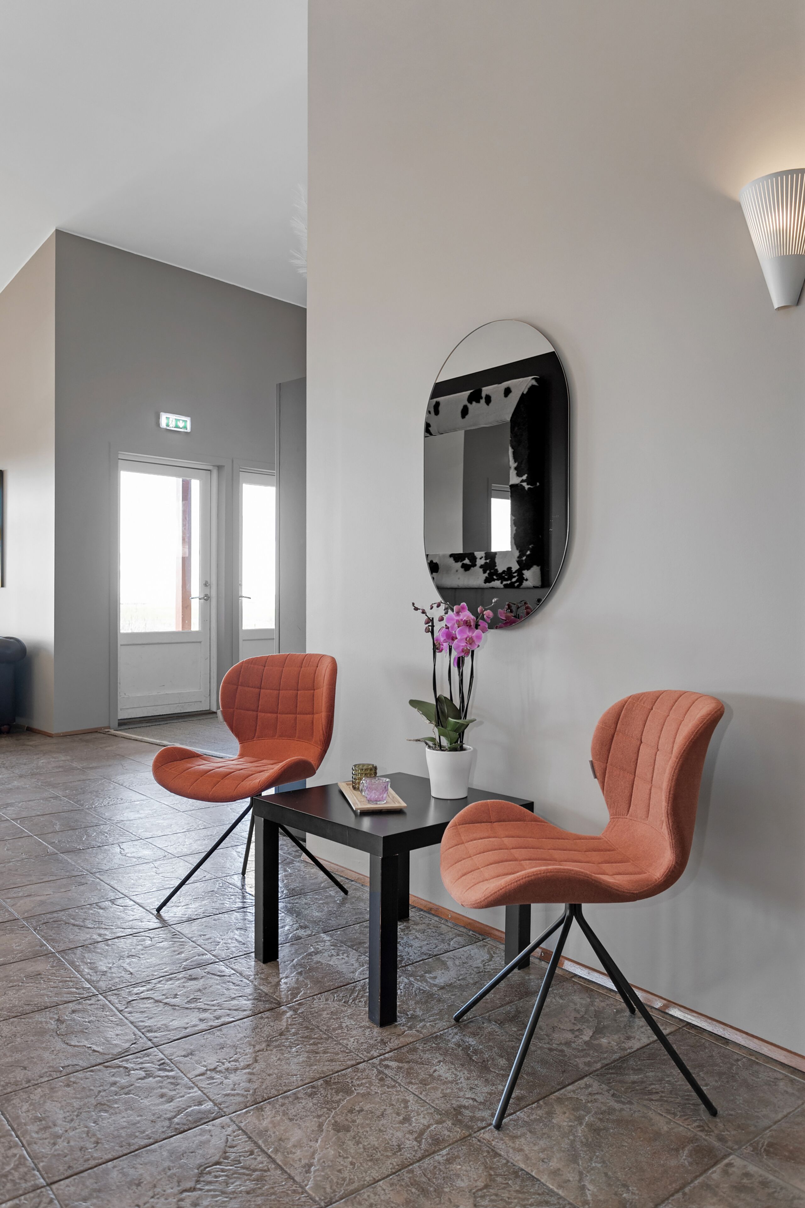 A modern interior with two orange chairs, a table, a potted orchid, and a large mirror on the wall.