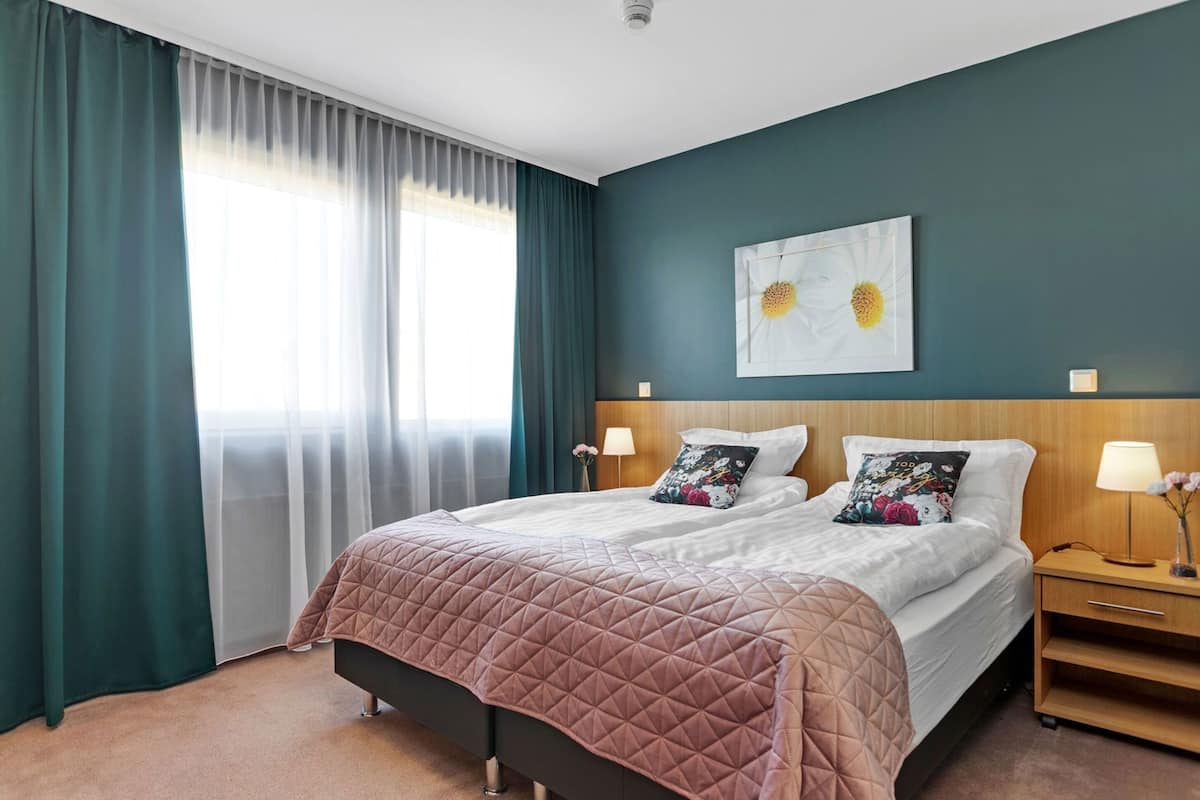 A double bed with a brown quilt, two bedside tables with lamps, a wall art of sunflowers, and a window with sheer curtains.