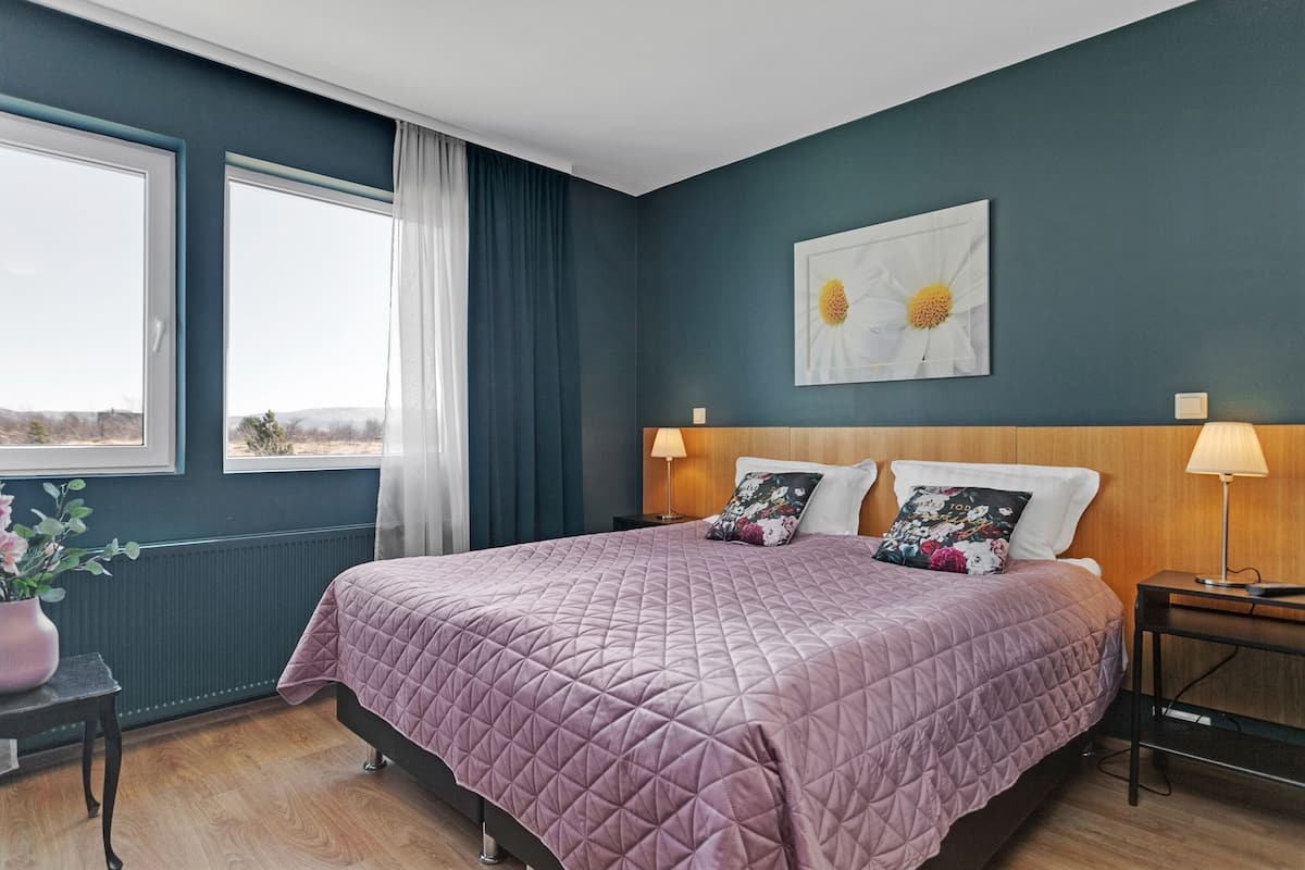 A bedroom with a large bed, two bedside lamps, a window with curtains, a wall-mounted flower painting, and a small table with a vase.