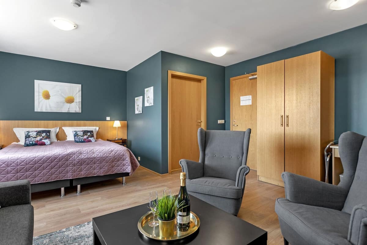 A modern bedroom with a bed, two armchairs, a wooden wardrobe, and a small table with a bottle and glasses.