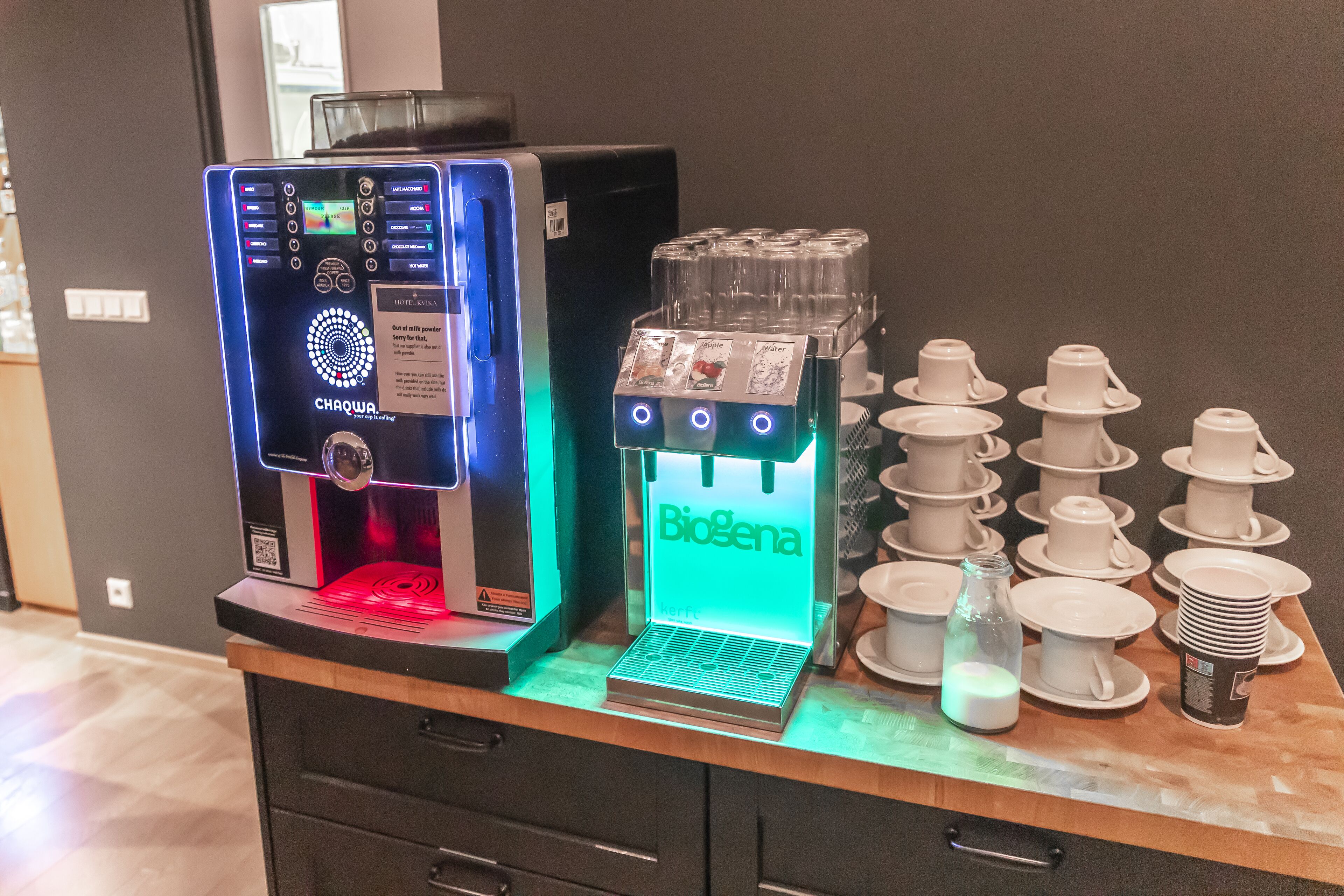 A coffee machine and a water dispenser on a counter with stacks of white cups and saucers.