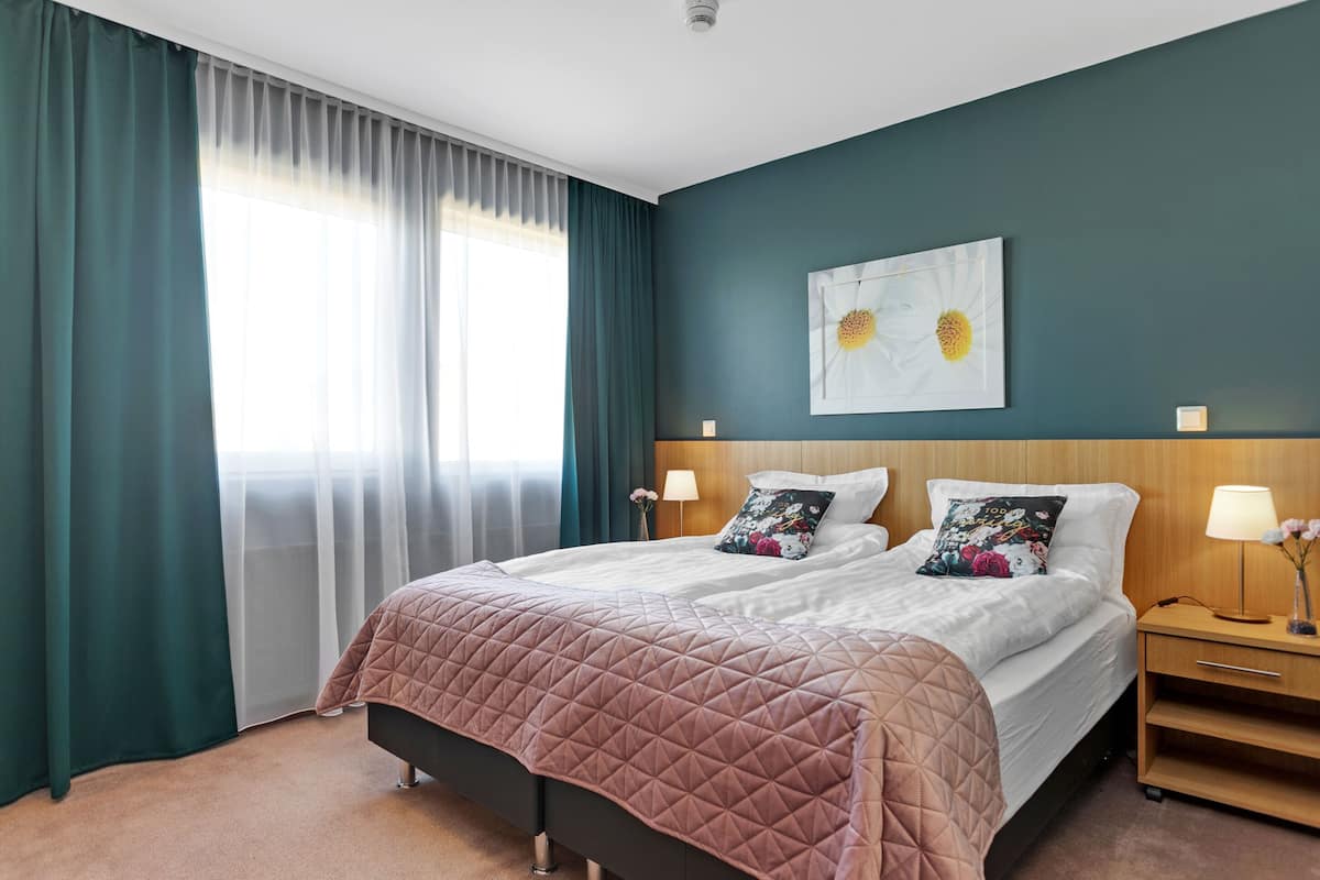 A double bed with a pink quilt, two bedside tables with lamps, a wall-mounted artwork, and a window with sheer curtains.