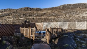 Un sentier en bois mène à un bâtiment doté d’un toit en pente, entouré de gros rochers et d’un paysage vallonné.