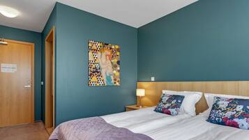 Habitación de hotel con paredes azul verdosas, una cama grande con ropa blanca, dos almohadones decorativos, un cabecero de madera, una mesita de noche con una lámpara y una obra de arte colorida en la pared.