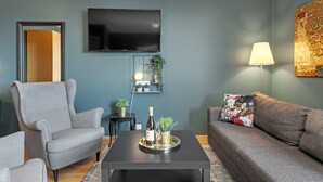 Un salon avec un fauteuil gris, un canapé, une table basse avec une bouteille et des verres, un téléviseur, une lampe et un élément décoratif mural.