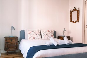 A neatly made bed with floral pillows, a blue blanket, and rolled towels. There are two bedside tables with lamps, a wooden dresser, and a mirror on the wall. - Casa Didi - Sintra (Sintra)
