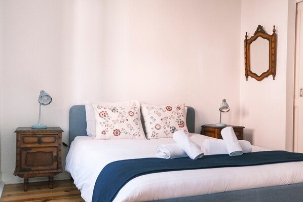 A neatly made bed with floral pillows, a blue blanket, and rolled towels. There are two bedside tables with lamps, a wooden dresser, and a mirror on the wall. - Casa Didi - Sintra (Sintra)
