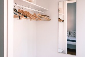 Una habitación con un armario blanco, perchas de madera y un espejo que refleja una cama con almohadas estampadas.