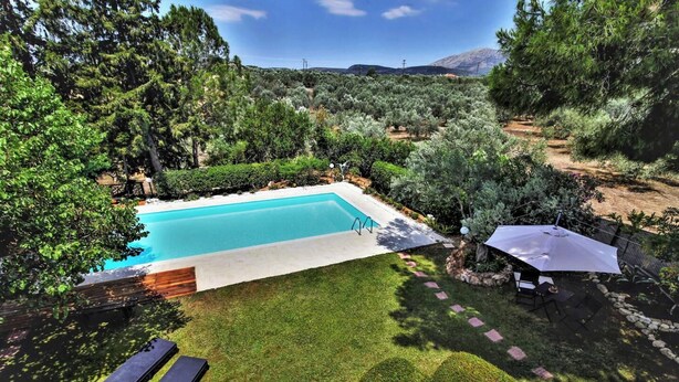 Una piscina rectangular rodeada de césped y setos bien cuidados, con un área de patio que incluye una mesa y sillas.