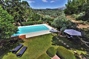 Une piscine rectangulaire entourée d’une pelouse et de haies bien entretenues, avec un espace patio comprenant une table et des chaises.