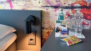 A hotel room with a bed, a bedside table with a bottle, glasses, a menu, and a packet of snacks. - Felix Hotel Darmstadt (Darmstadt)