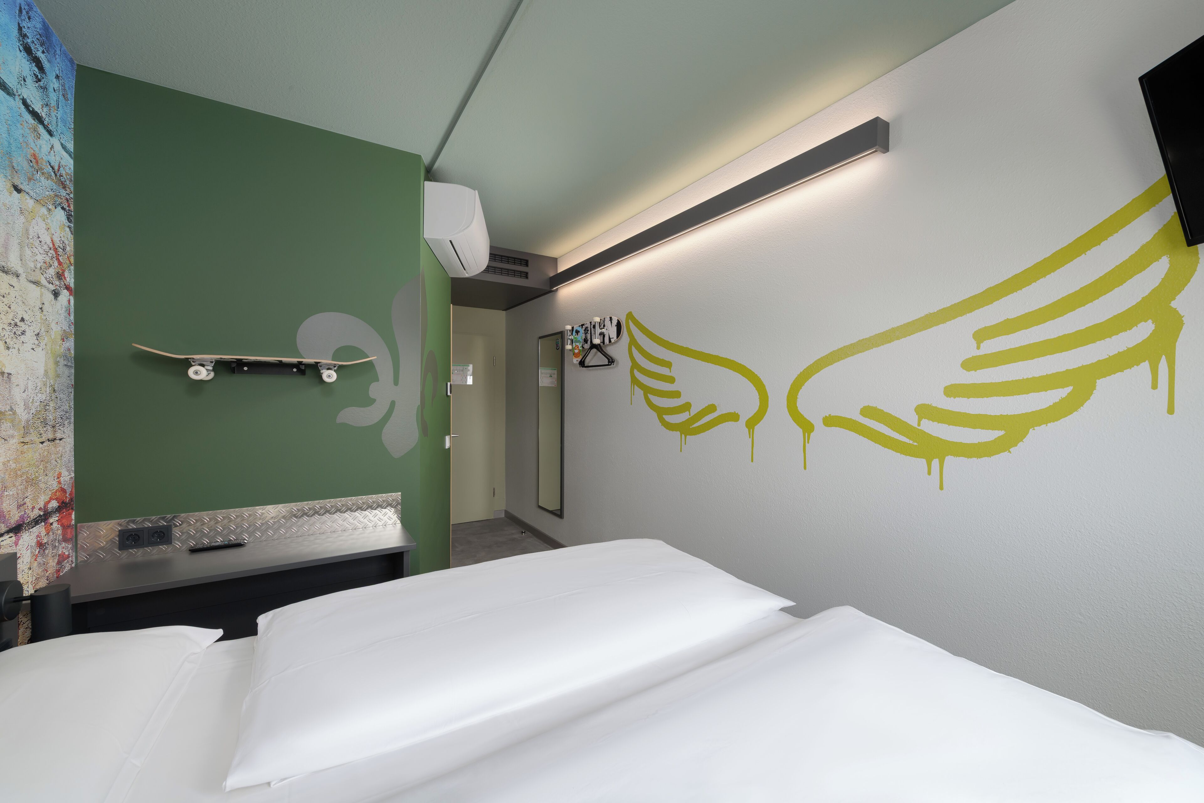 Hotelkamer met een bed, een wandairconditioner, een spiegel en een muur met een gele vleugelprint.