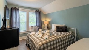 A bedroom with a checkered bedspread, a bedside lamp, a window with curtains, a desk with a computer, and a chair. - Room 8 (3rd Floor and Shared Bathroom) (Laconia)