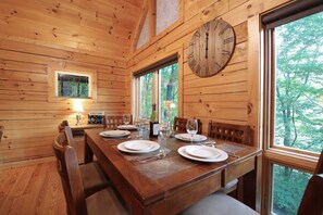 A wooden dining room with a large wooden clock, a window with a view of trees, and a table set for four. - Bearadise Mtn Cabin~Hot Tub+Fire Pit, Grill & WiFi (Burnsville)