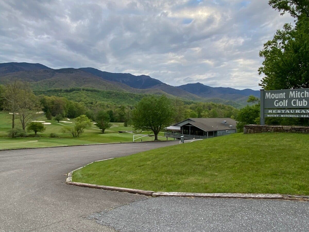 A golf club with a building and a sign, surrounded by a green landscape and mountains.
