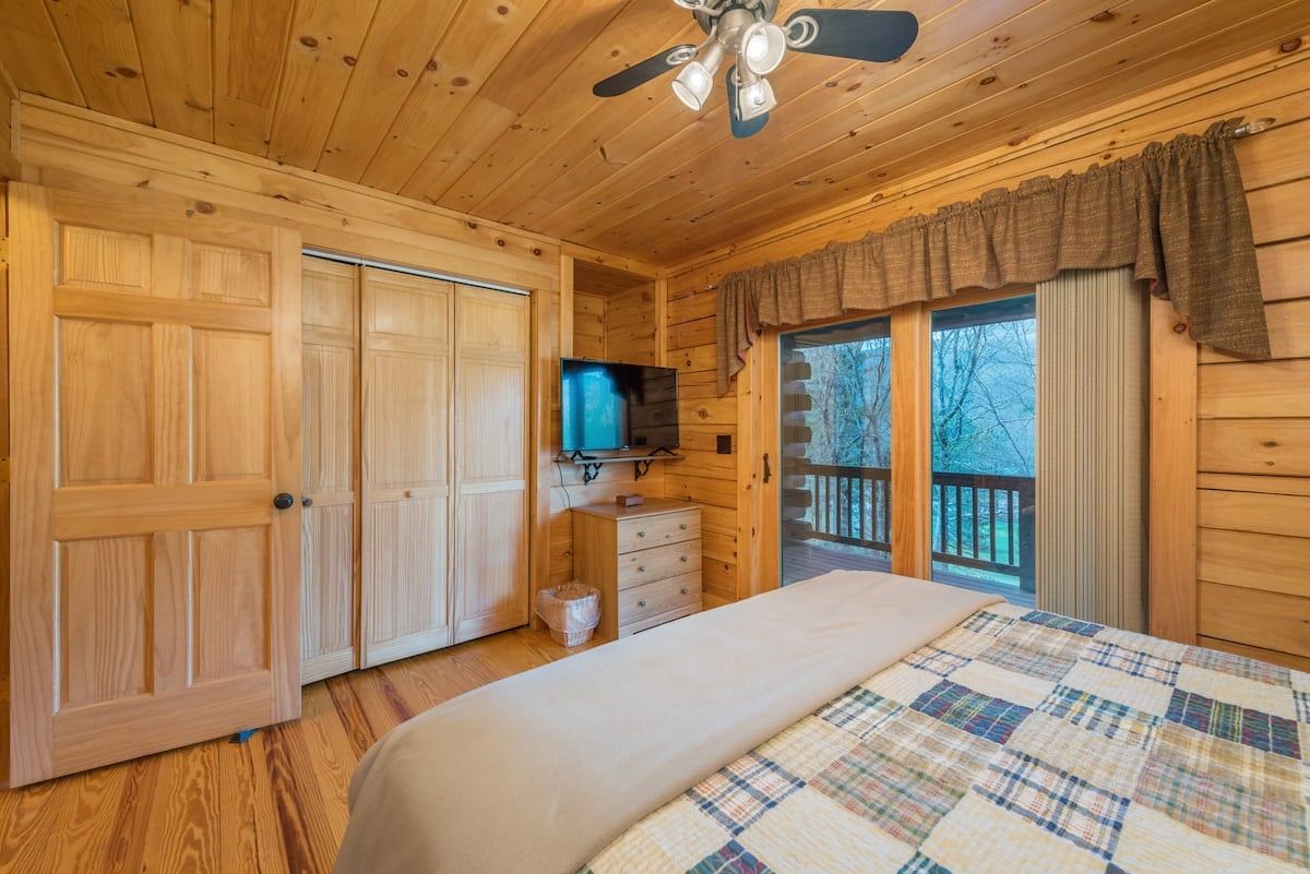 A wooden cabin room with a bed, a television, a dresser, and a sliding glass door leading to a balcony.