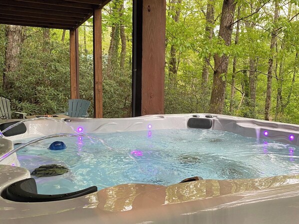 A hot tub with water jets and LED lights, surrounded by a wooden deck and trees. - Bearadise Mtn Cabin~Hot Tub+Fire Pit, Grill & WiFi (Burnsville)