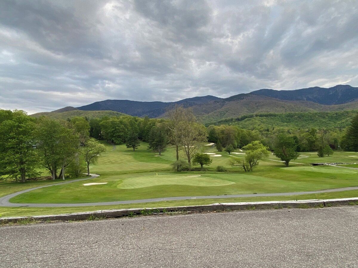Un campo de golf con una carretera en primer plano, árboles y montañas al fondo.