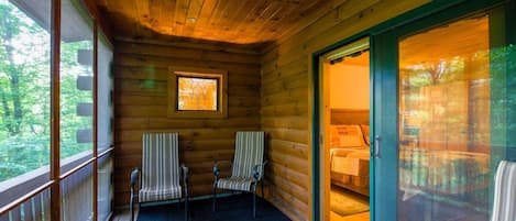 A cabin with a porch, two chairs, a window, and a sliding glass door leading to a bedroom.