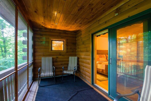 A cabin with a porch, two chairs, a window, and a sliding glass door leading to a bedroom.