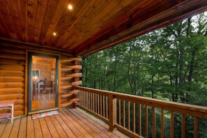 A log cabin with a wooden deck, a glass door leading to an interior dining area, and a view of a forested landscape. - Bearadise Mtn Cabin~Hot Tub+Fire Pit, Grill & WiFi (Burnsville)