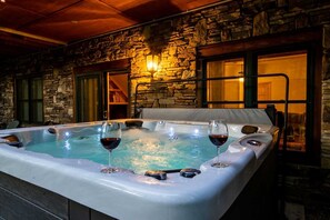 A hot tub with two glasses of red wine, set against a stone wall with a window and a door. - Bearadise Mtn Cabin~Hot Tub+Fire Pit, Grill & WiFi (Burnsville)