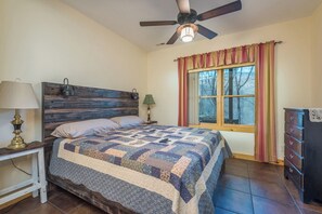 A bedroom with a wooden headboard, a quilted bedspread, a nightstand with a lamp, a dresser, and a window with curtains.