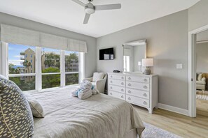 Un dormitorio con cama, cómoda, ventilador de techo y ventana con vistas a edificios y árboles.