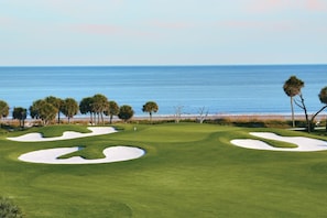 A golf course with sand bunkers and palm trees, overlooking a beach and ocean.