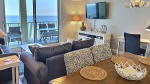 A living room with a wooden dining table, dark sofas, a TV, and a balcony with a view of the ocean. - TOP RATED / HUGE Balcony Beachfront 3 bed / 3 bath! (Panama City Beach)
