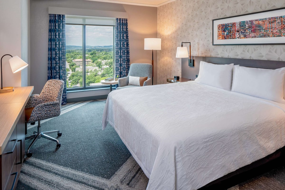 Un cuarto de hotel con una cama grande, un escritorio con lámpara, una silla, una mesita y una ventana con vista a la ciudad.
