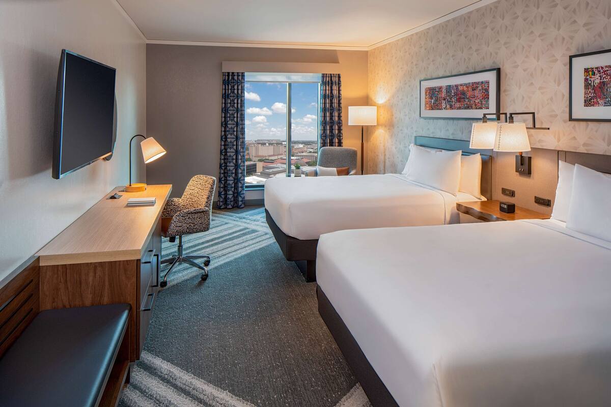 Habitación de hotel con dos camas, un escritorio, una silla, un televisor y vistas a la ciudad.