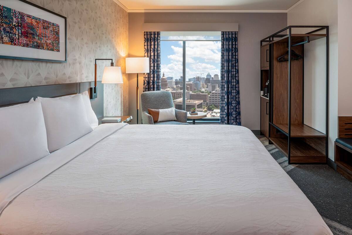 Habitación de hotel con una cama grande, una silla, un escritorio y un armario. Se ve la ciudad desde la ventana.