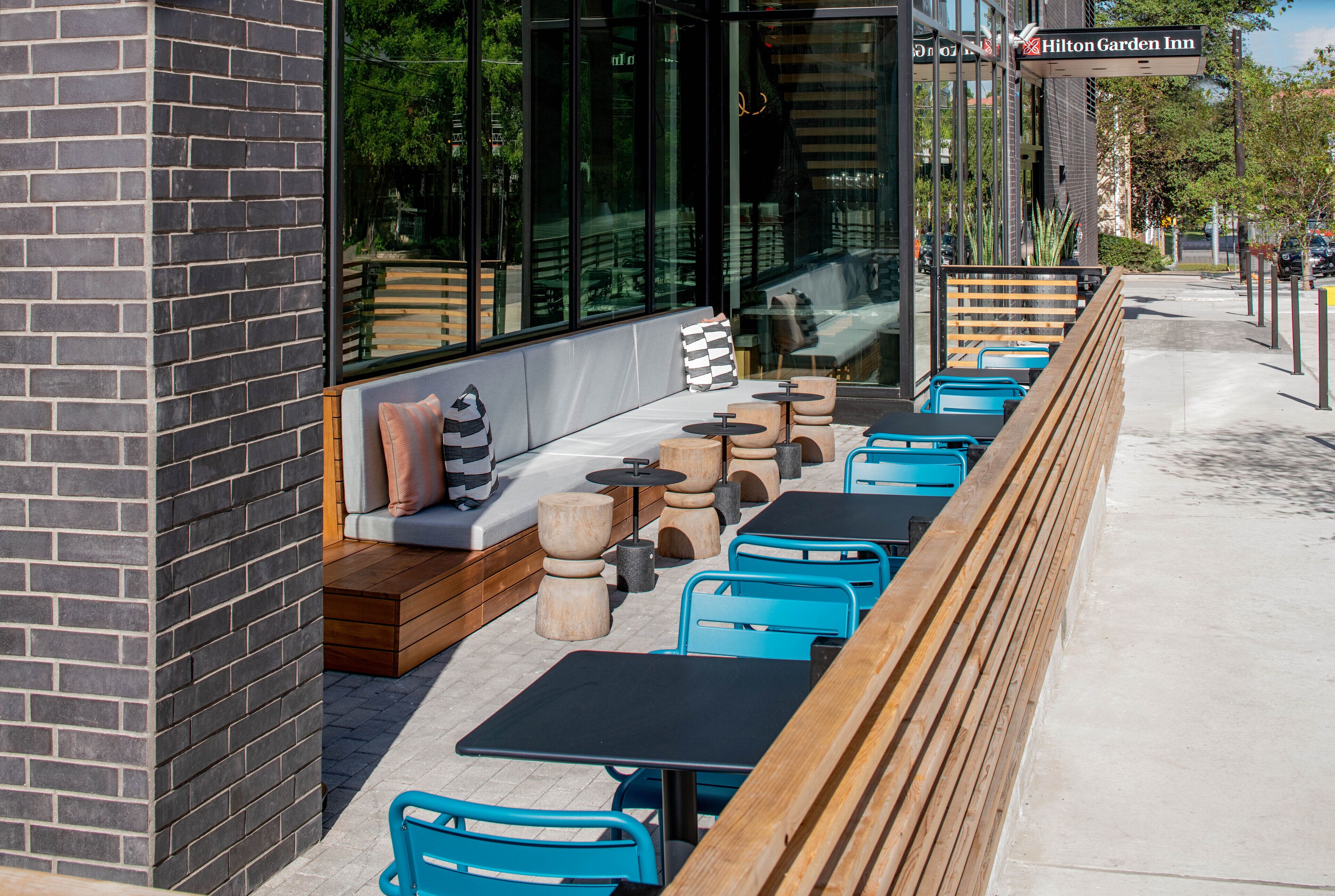 An outdoor seating area with wooden benches and blue chairs, a brick wall, and a sign for Hilton Garden Inn.