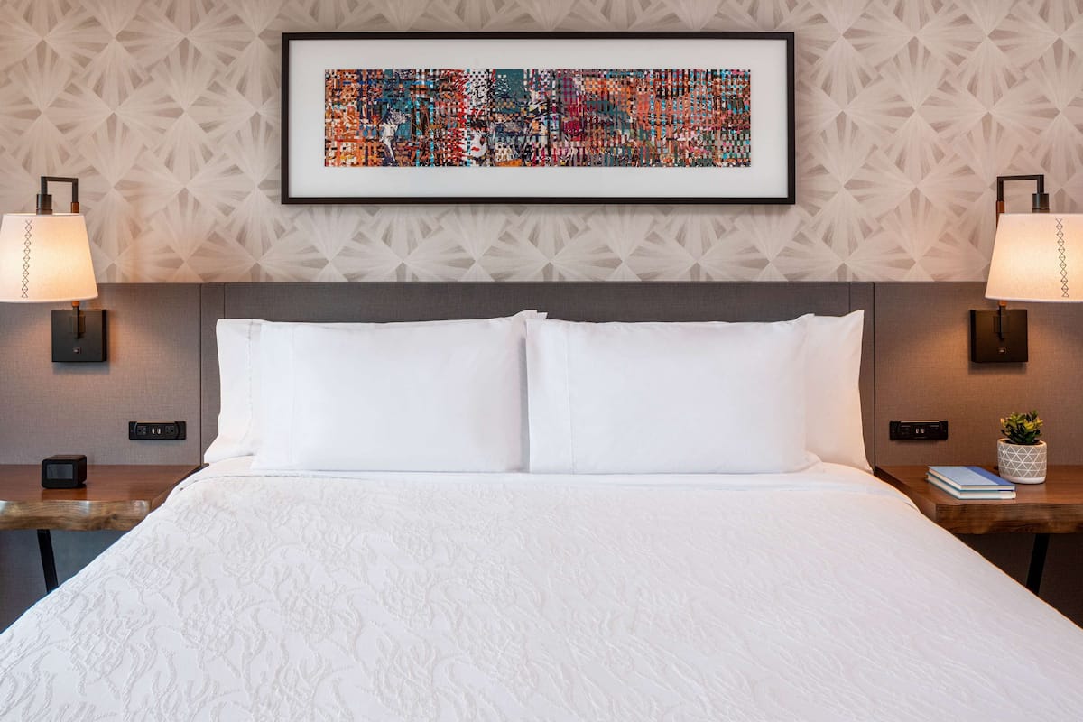 Habitación de hotel con una cama grande, dos mesitas de noche, un cuadro abstracto enmarcado y dos lámparas de pared.