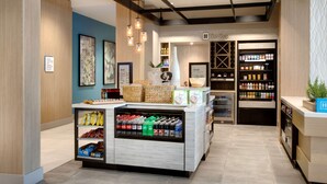 A modern retail area with a central counter displaying snacks and beverages, shelves with bottled drinks, and a section labeled 'The Shop' with various products.