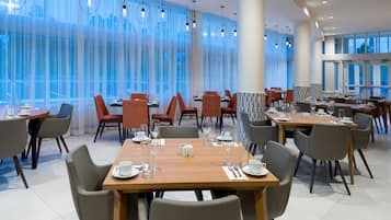 Un espace de restauration moderne, doté de plusieurs tables et chaises, de grandes fenêtres avec des rideaux légers et d’un éclairage suspendu.