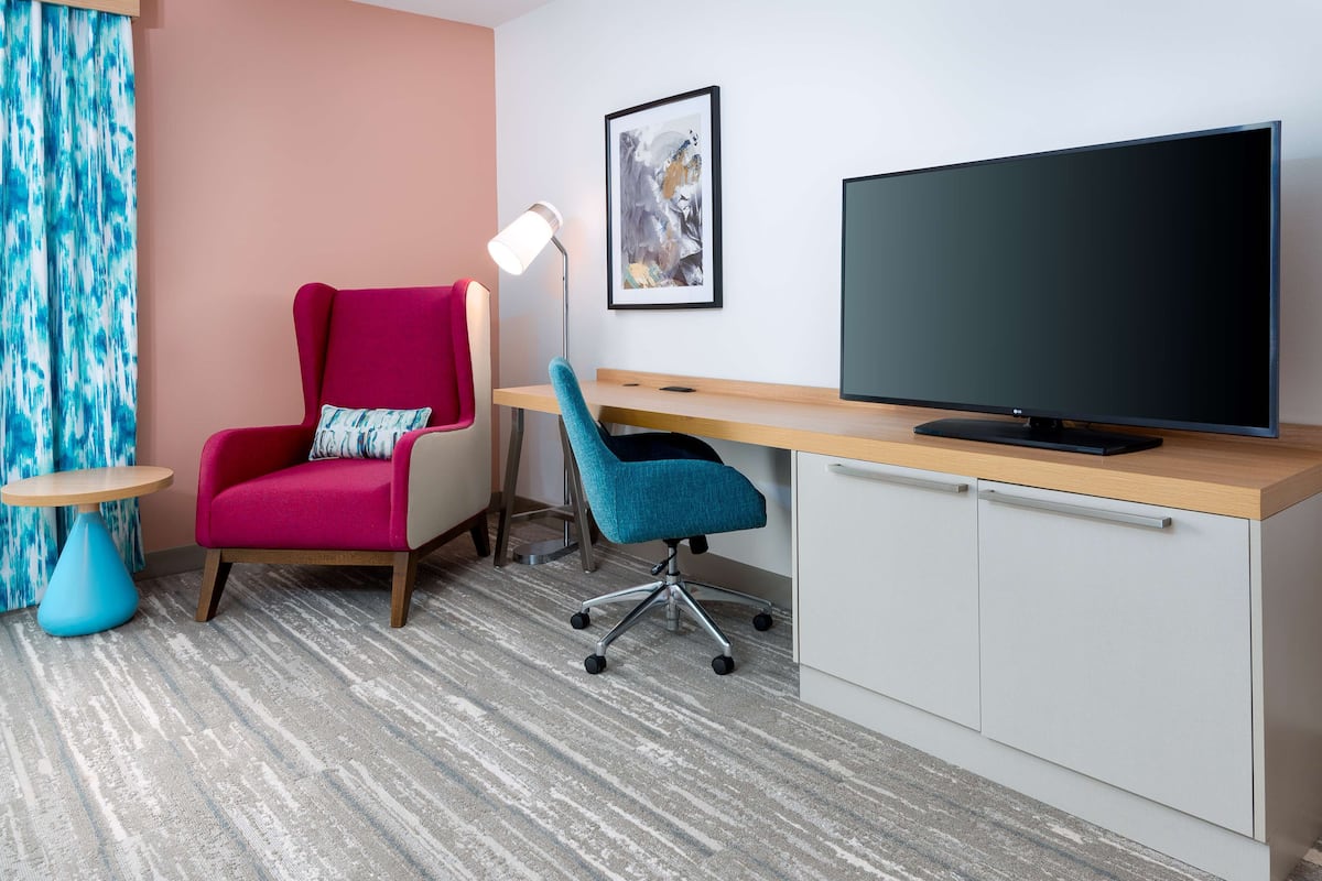 Une chambre d’hôtel moderne avec un mur d’accent rose, un grand téléviseur à écran plat, un bureau avec un ordinateur, une chaise de bureau bleue, une petite table ronde et un rideau bleu.