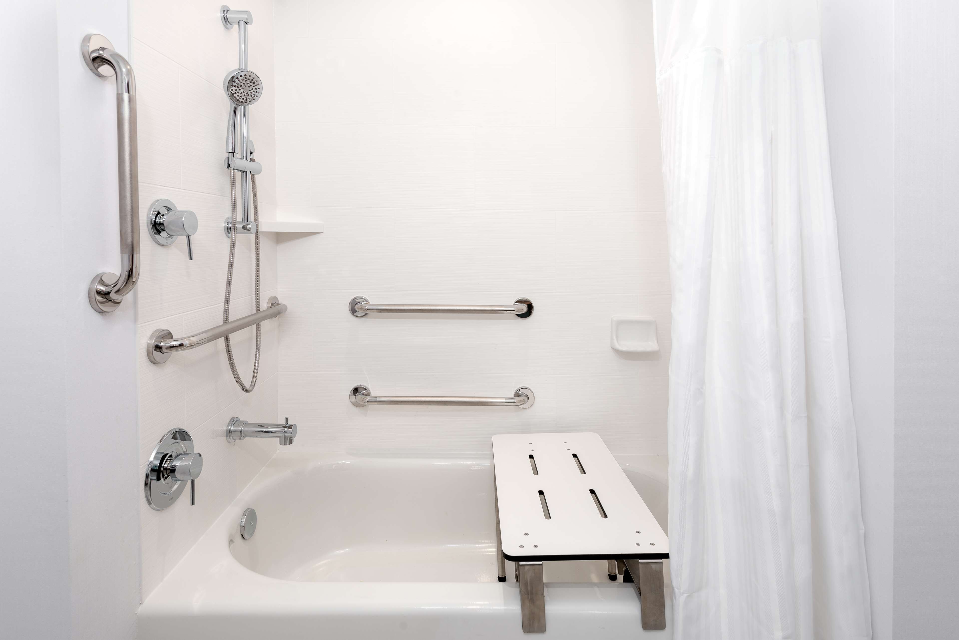 Un baño con bañera, barras de apoyo, cabezal de ducha y una cortina blanca.