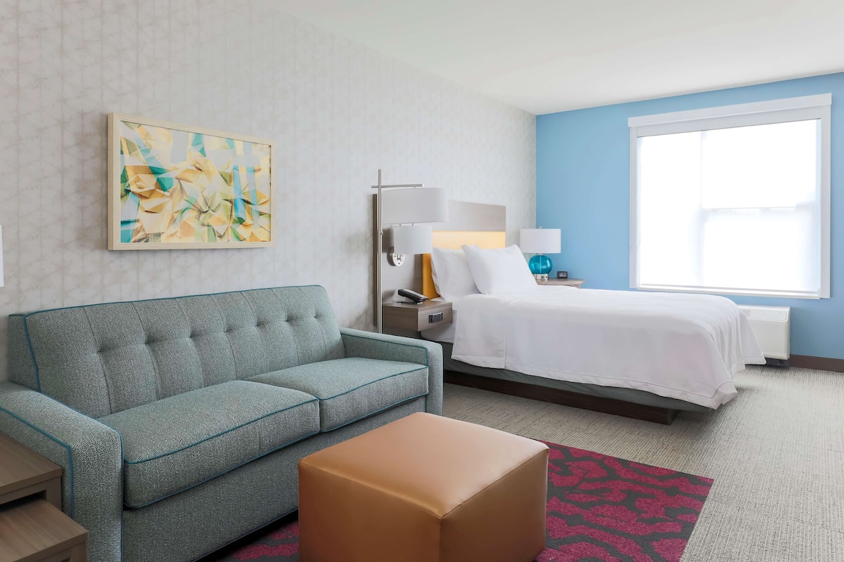 A hotel room with a sofa, a bed, a nightstand, a lamp, and a painting on the wall.