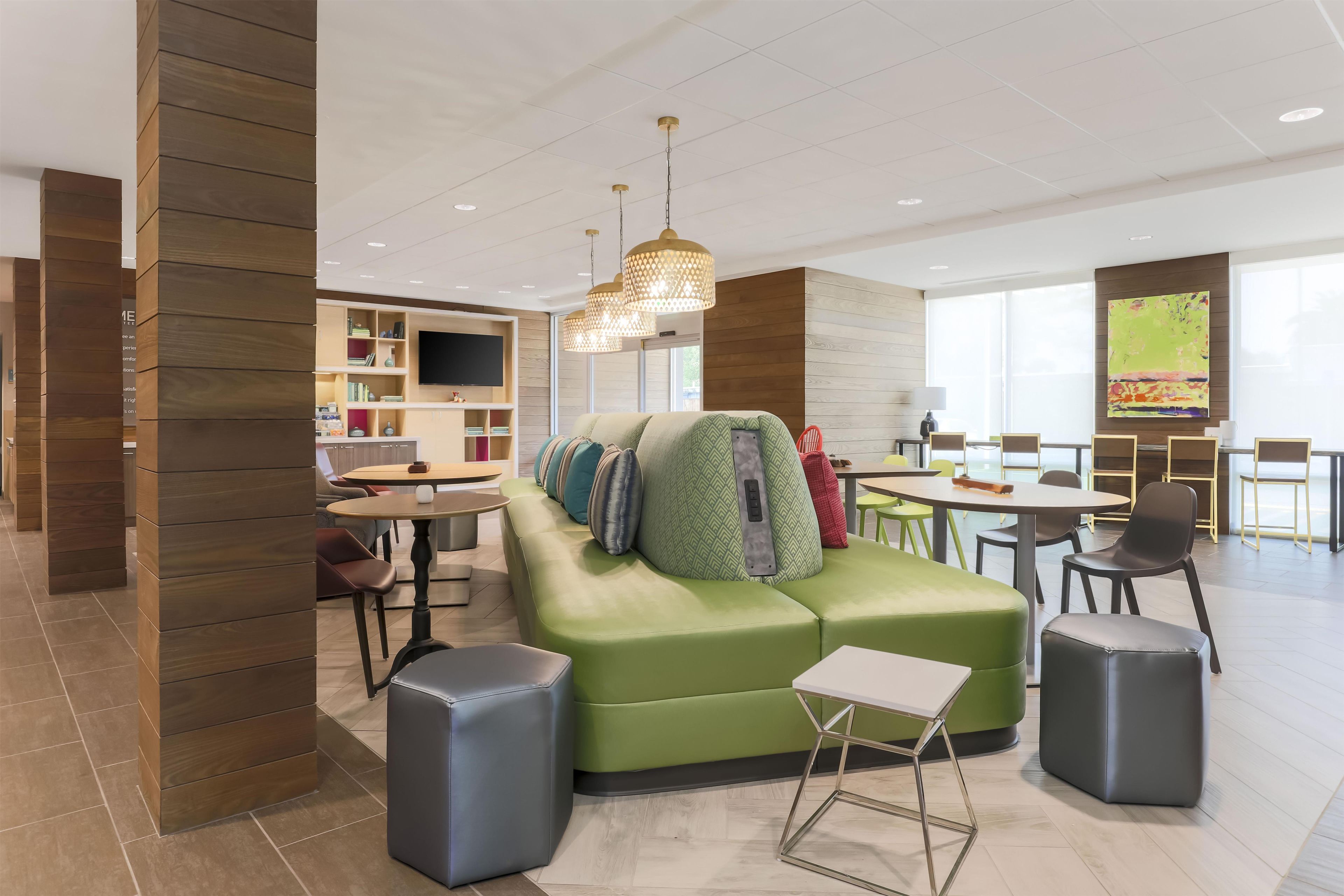 Ein moderner Lounge-Bereich mit einem grünen Sofa, verschiedenen Sitzgelegenheiten, einem Fernseher und einem großen Fenster, das viel Tageslicht hereinlässt.