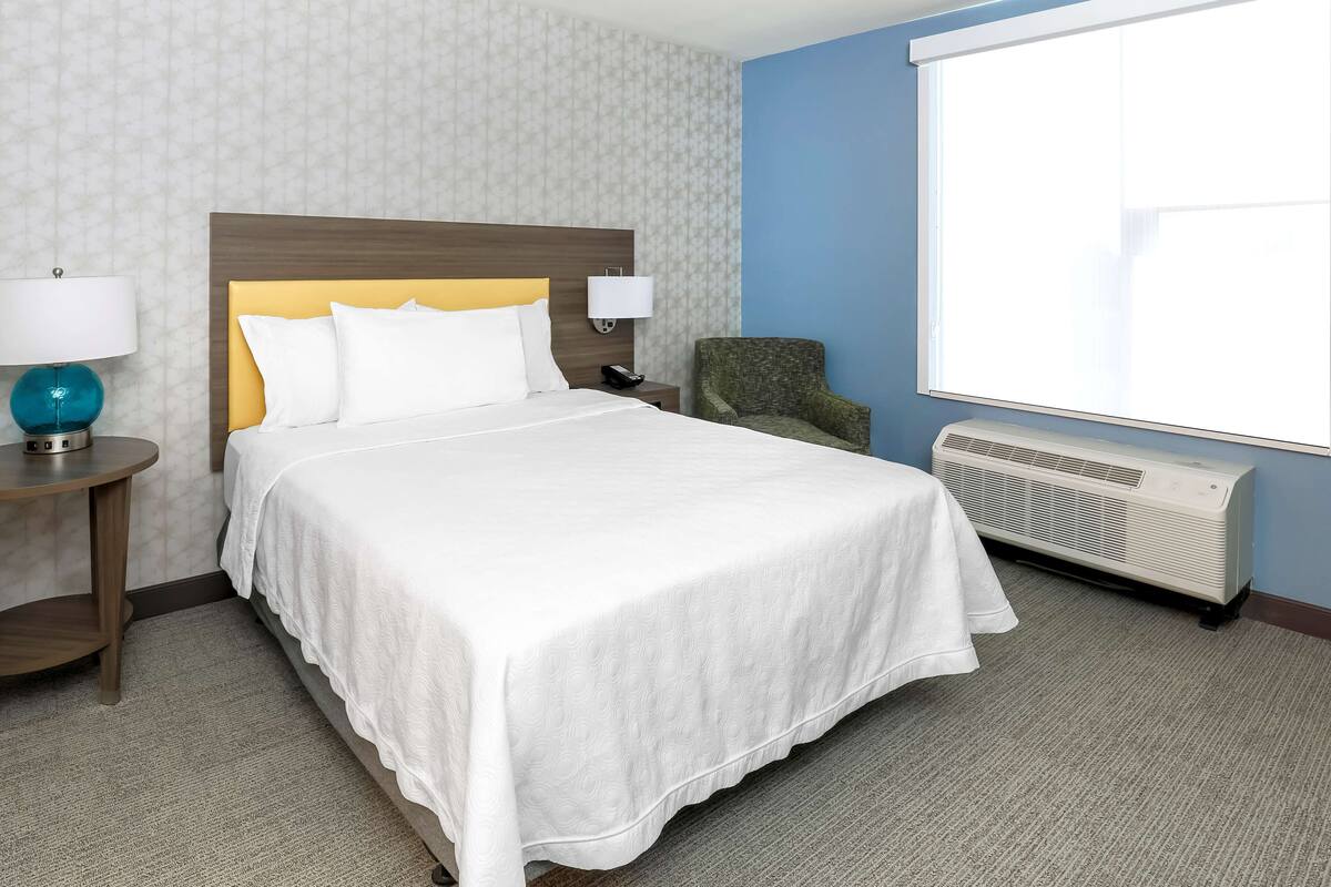 A hotel room with a large bed, a nightstand with a lamp, a chair, a window, and an air conditioning unit.
