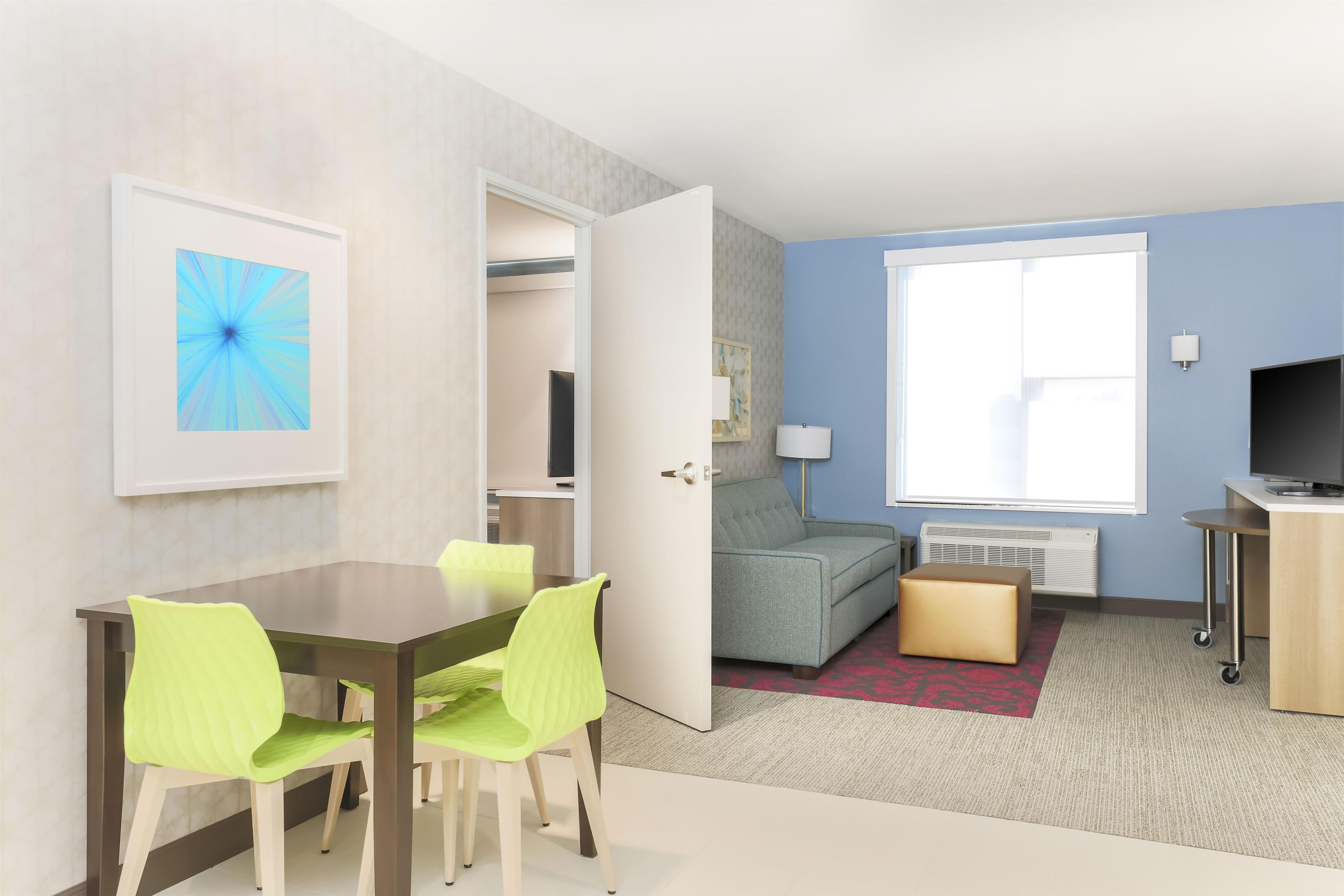 A modern hotel room with a dining area, a sofa, a TV, and a window with natural light.