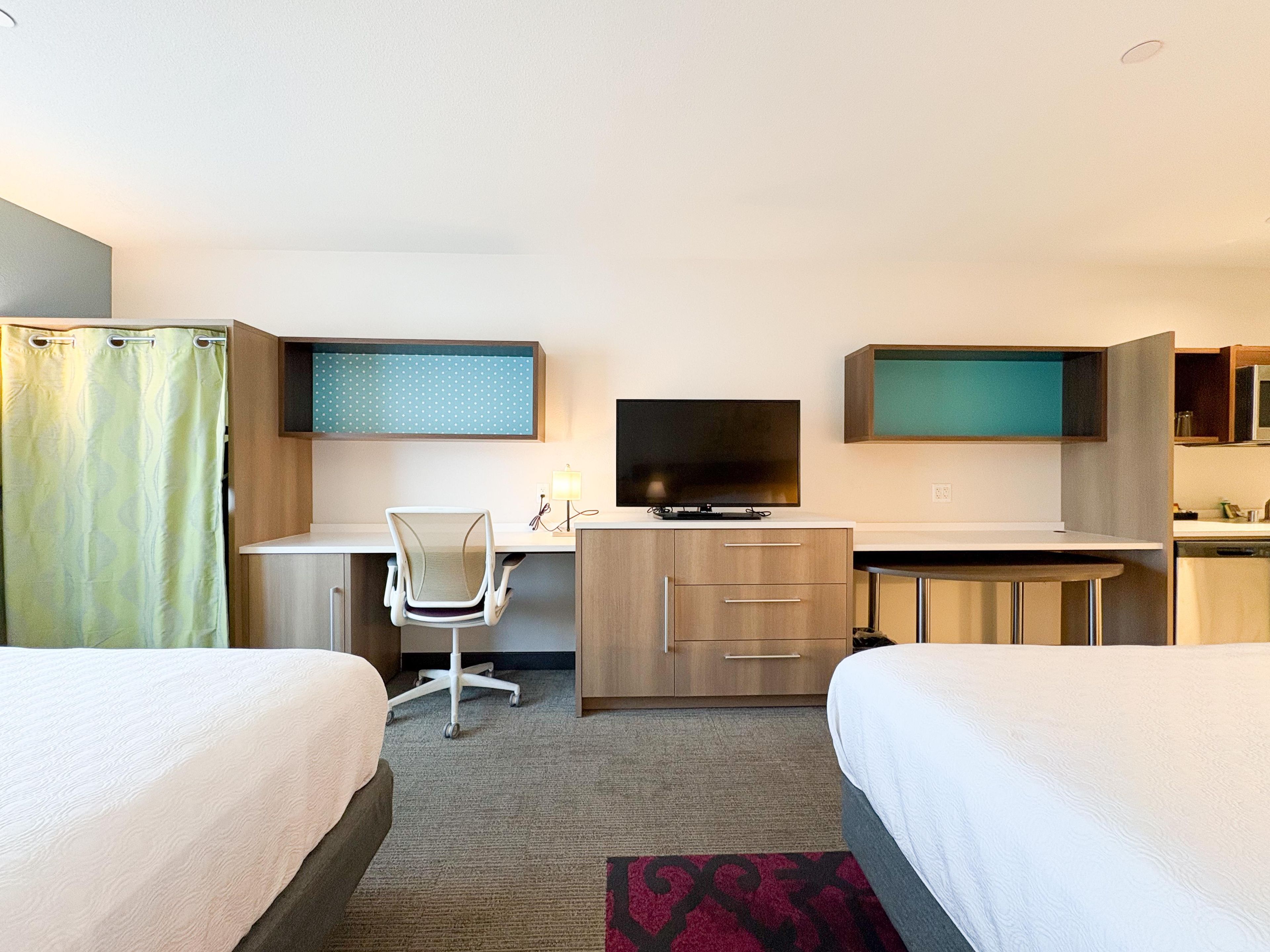 A hotel room with two beds, a desk with a chair, a television, and a small kitchenette.