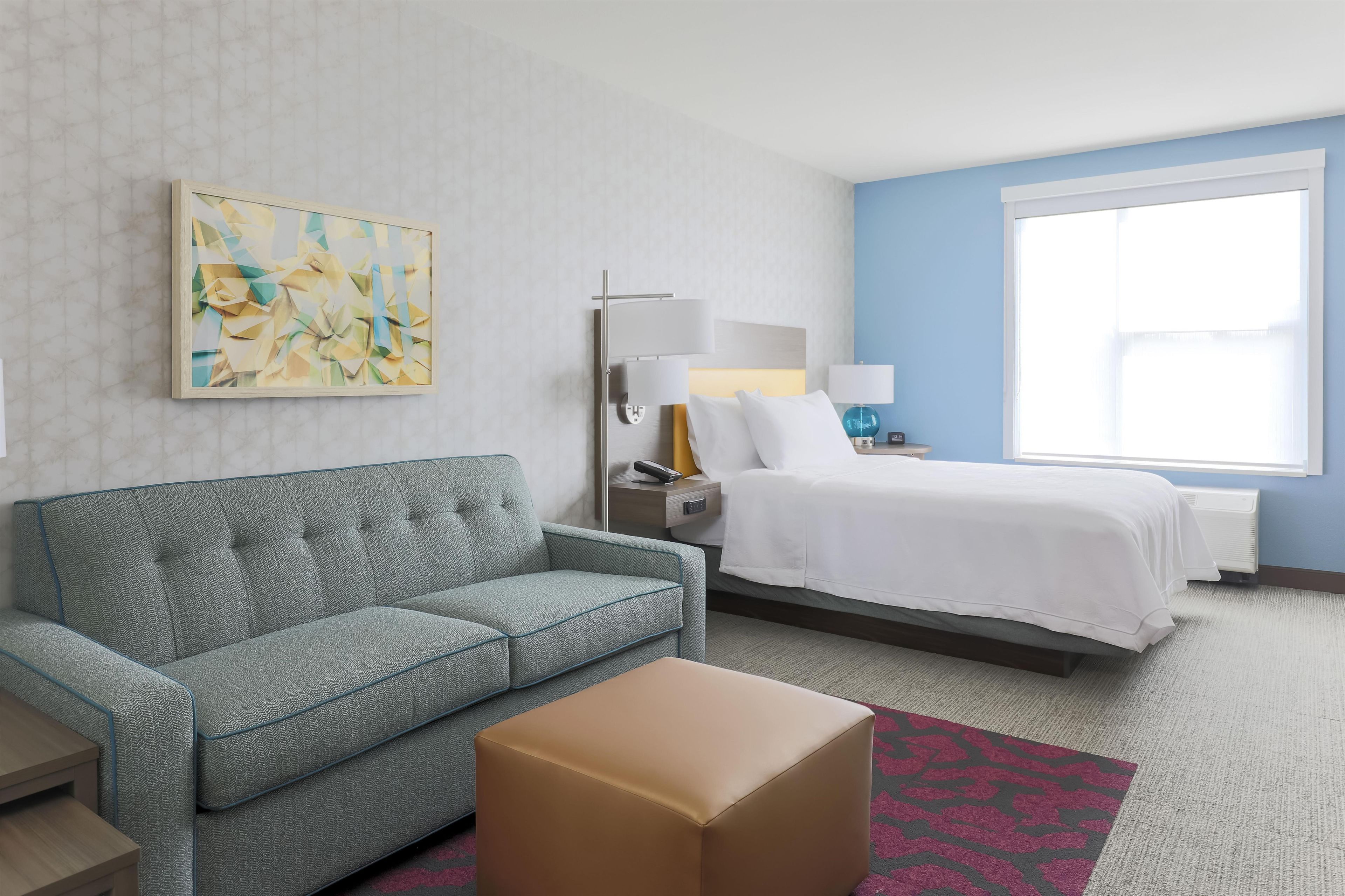 Une chambre d’hôtel avec un canapé, un lit, une table de chevet avec une lampe, une œuvre d’art accrochée au mur et une grande fenêtre.