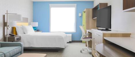 A hotel room with a bed, bedside table, lamp, television, desk, chair, and a window.