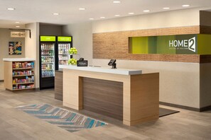 Een hotel receptiegedeelte met een receptiebalie, een minibar, een koelkast gevuld met drankjes en een wandbordje met 'HOME 2 Suites by Hilton'.