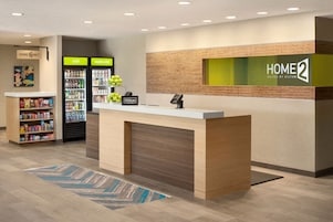 Zona de recepción de un hotel con mostrador de recepción, un minibar, un refrigerador lleno de bebidas y un letrero en la pared que dice 'HOME 2 Suites by Hilton'.