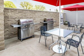 Un patio extérieur équipé de deux barbecues Char-Broil en acier inoxydable, d’une table, de chaises et d’un parasol rouge.